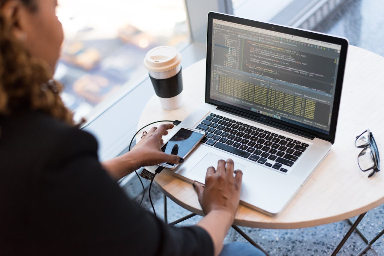 process-img Black woman programming on a laptop with coffee, smartphone, and glasses on a desk in an office.